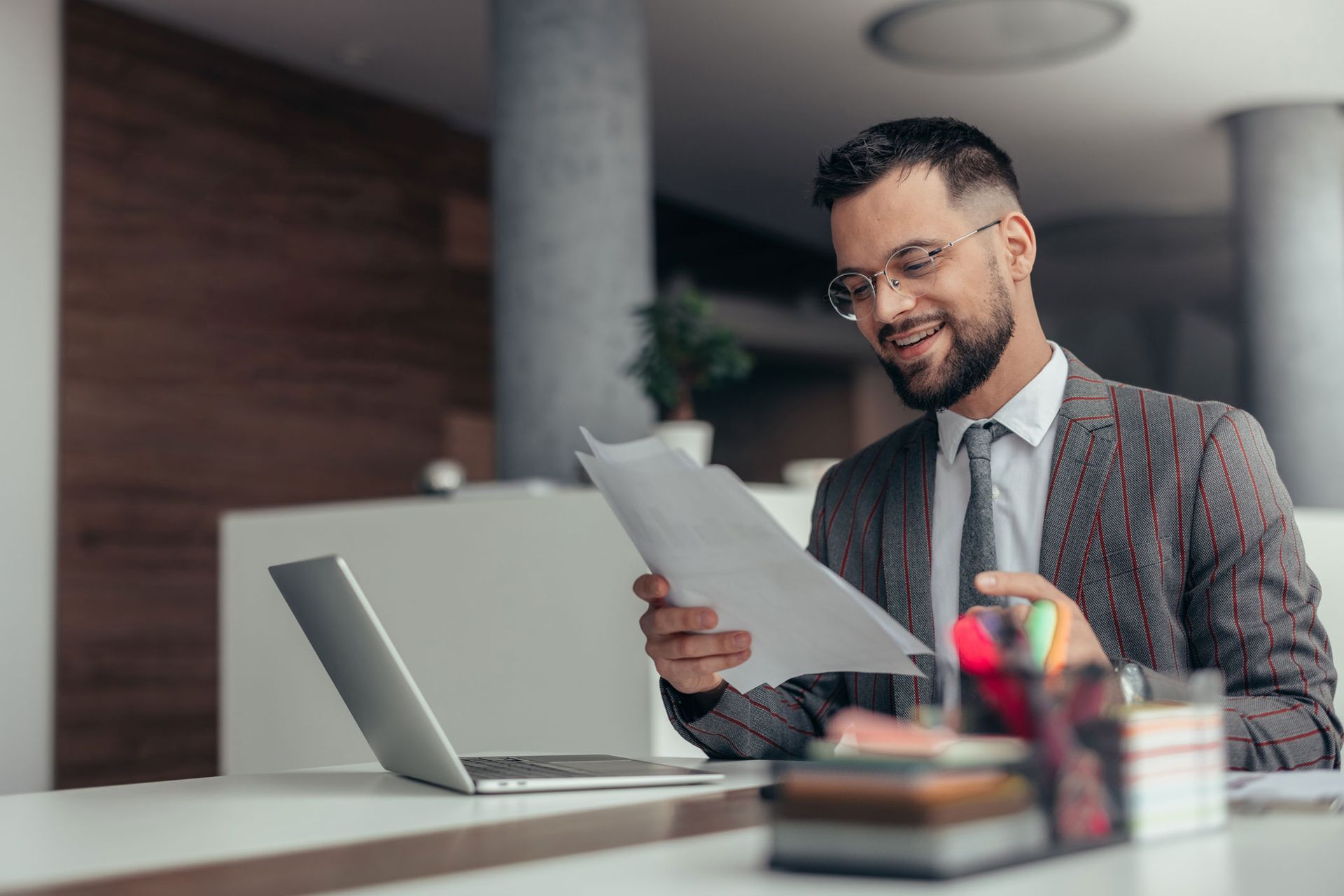 A man is sitting at a desk in front of a laptop computer holding a piece of paper.