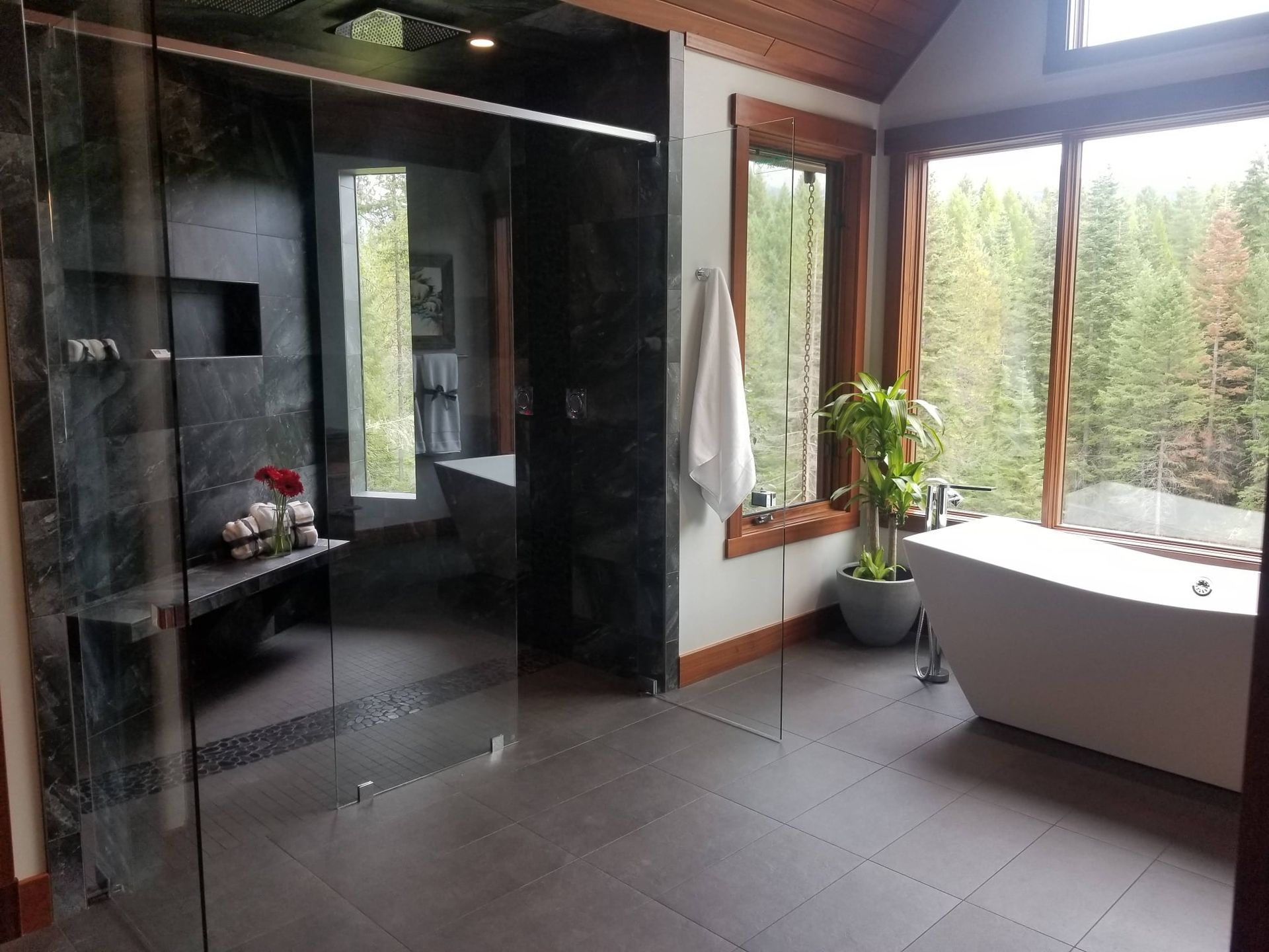Luxurious bathroom with large windows overlooking a forest, featuring a shower, tub, and tiled floor.