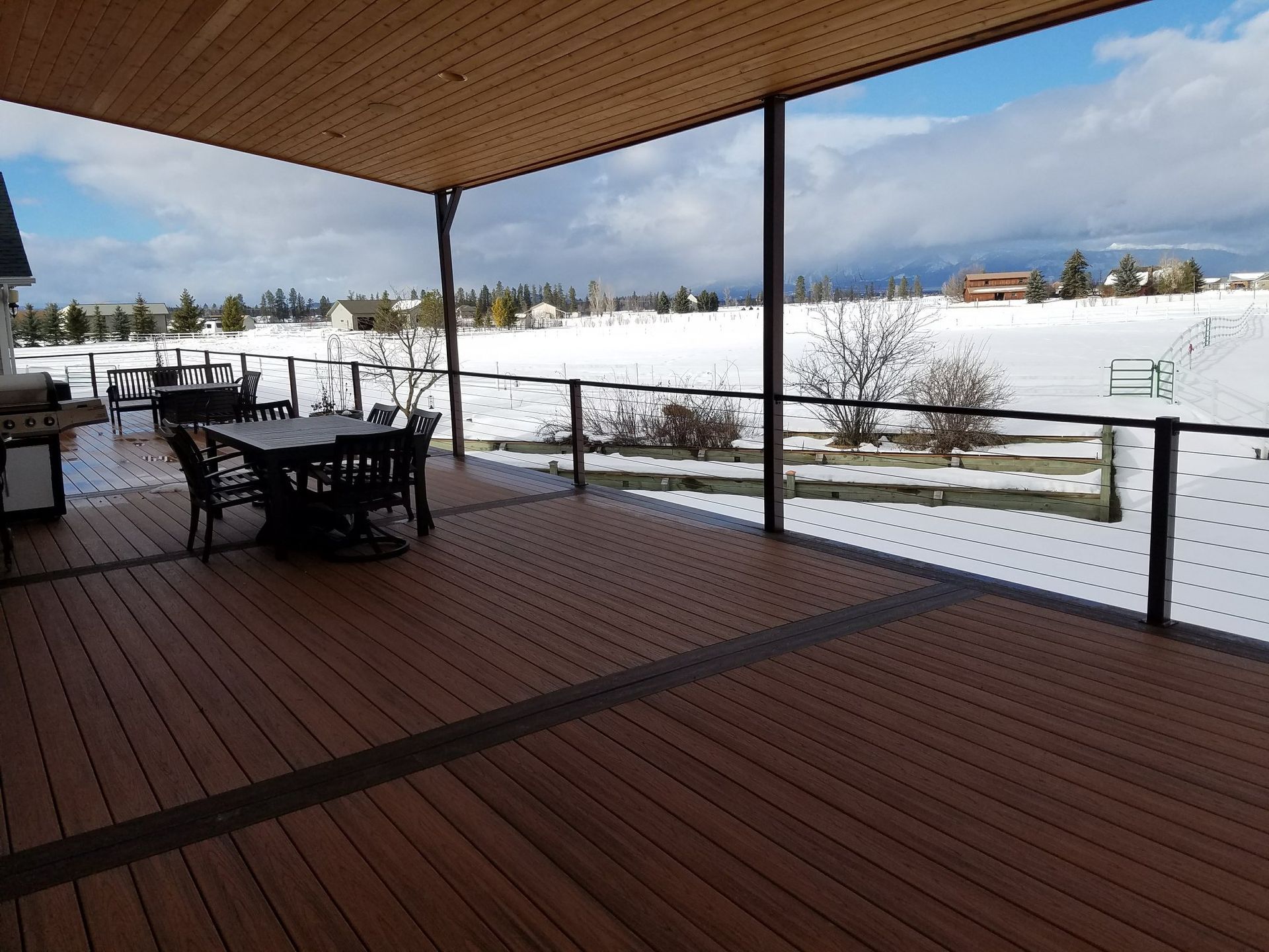 A spacious wooden deck with dining table and grill overlooking a snowy field and distant buildings under a partly cloudy sky.