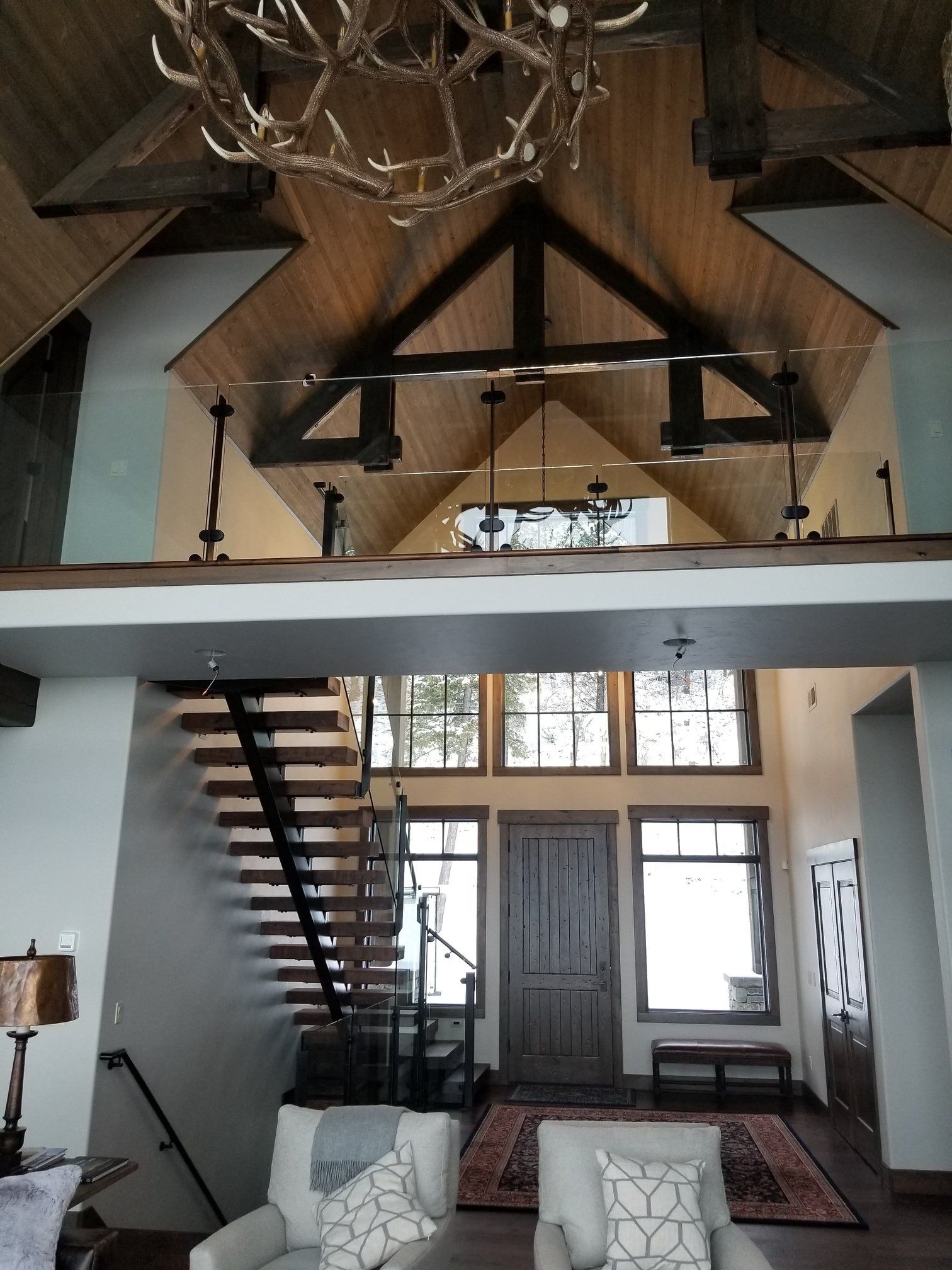 Lodge interior with wooden beams, staircase, and chandelier. Sunlight through windows, chairs in foreground.