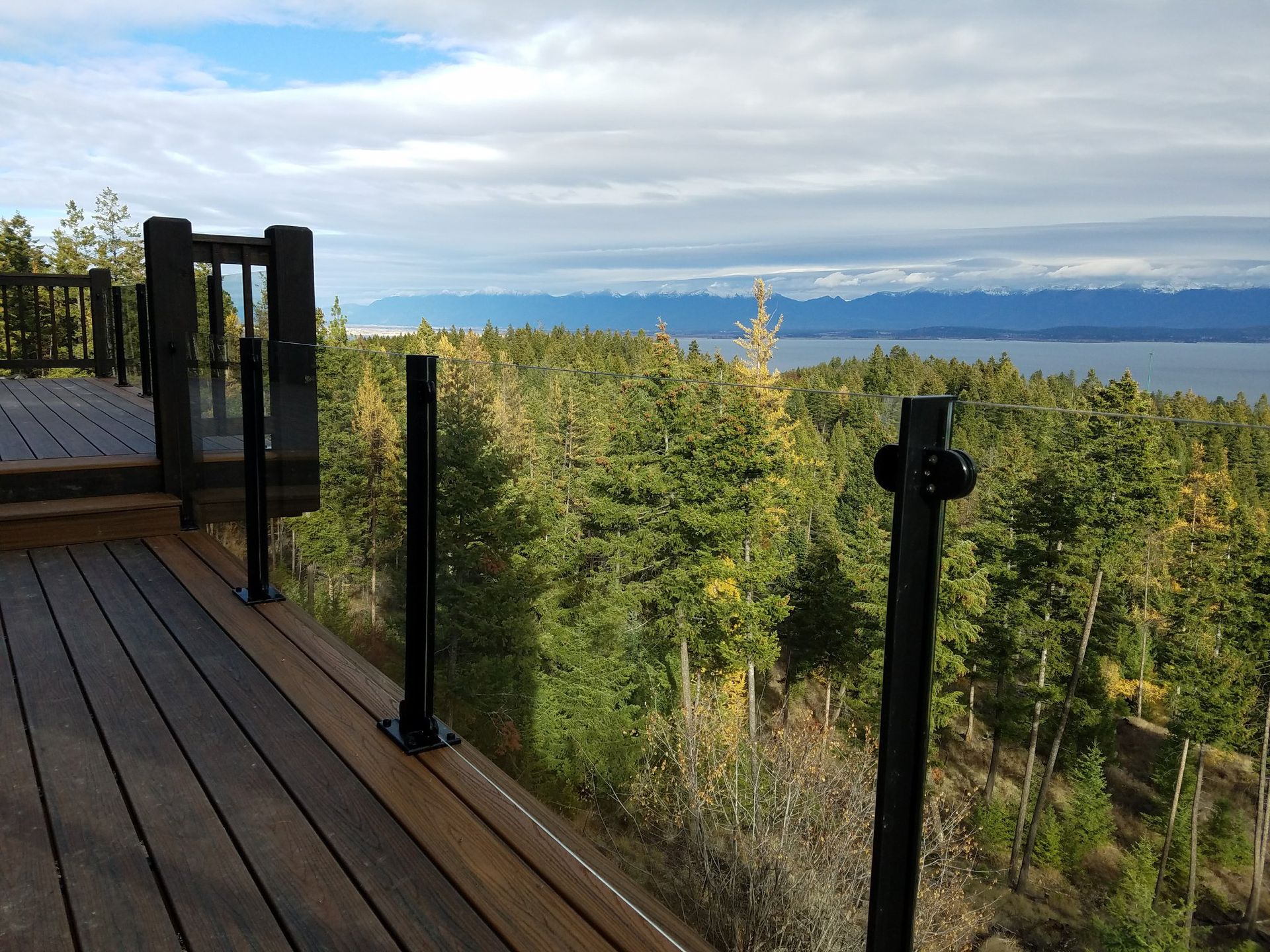 Wooden deck with black railings overlooking a forest and mountains under a cloudy sky.