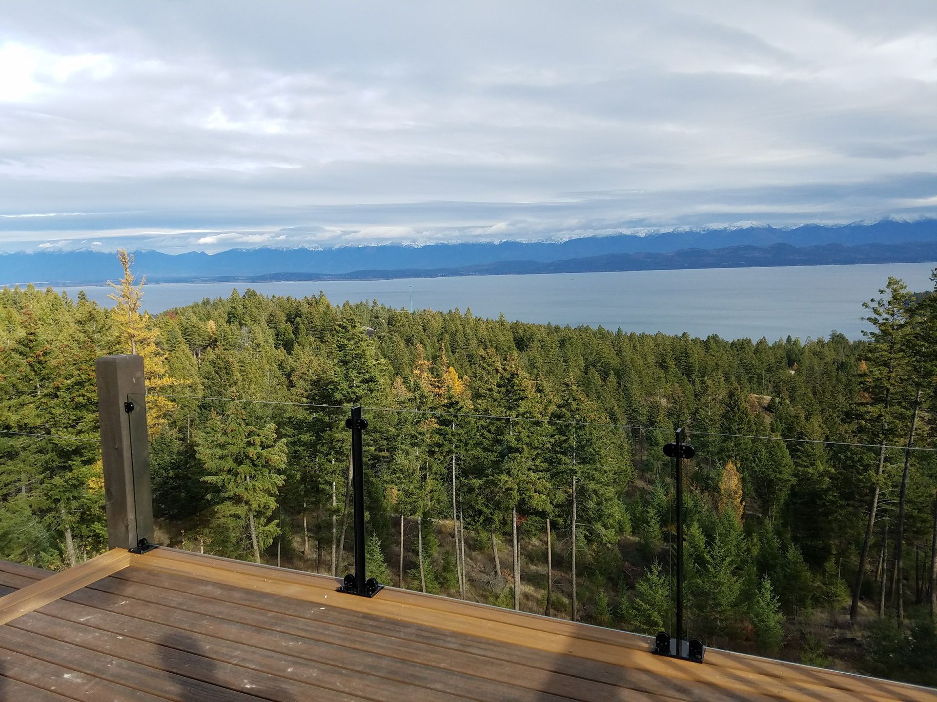 View from a wooden deck overlooking a forest, lake, and distant mountains under a cloudy sky.