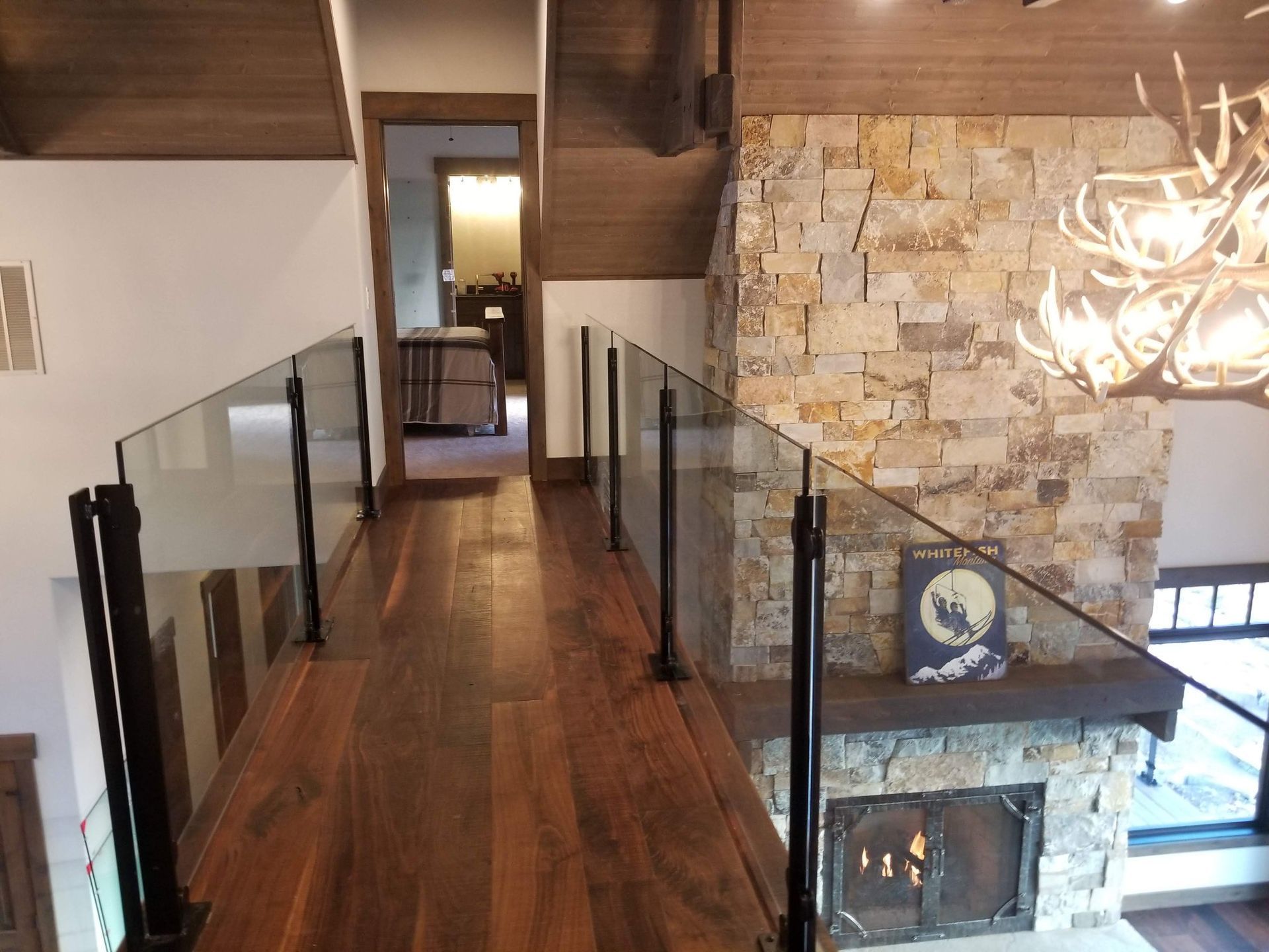 Wooden walkway with glass railings, stone fireplace, and a chandelier.