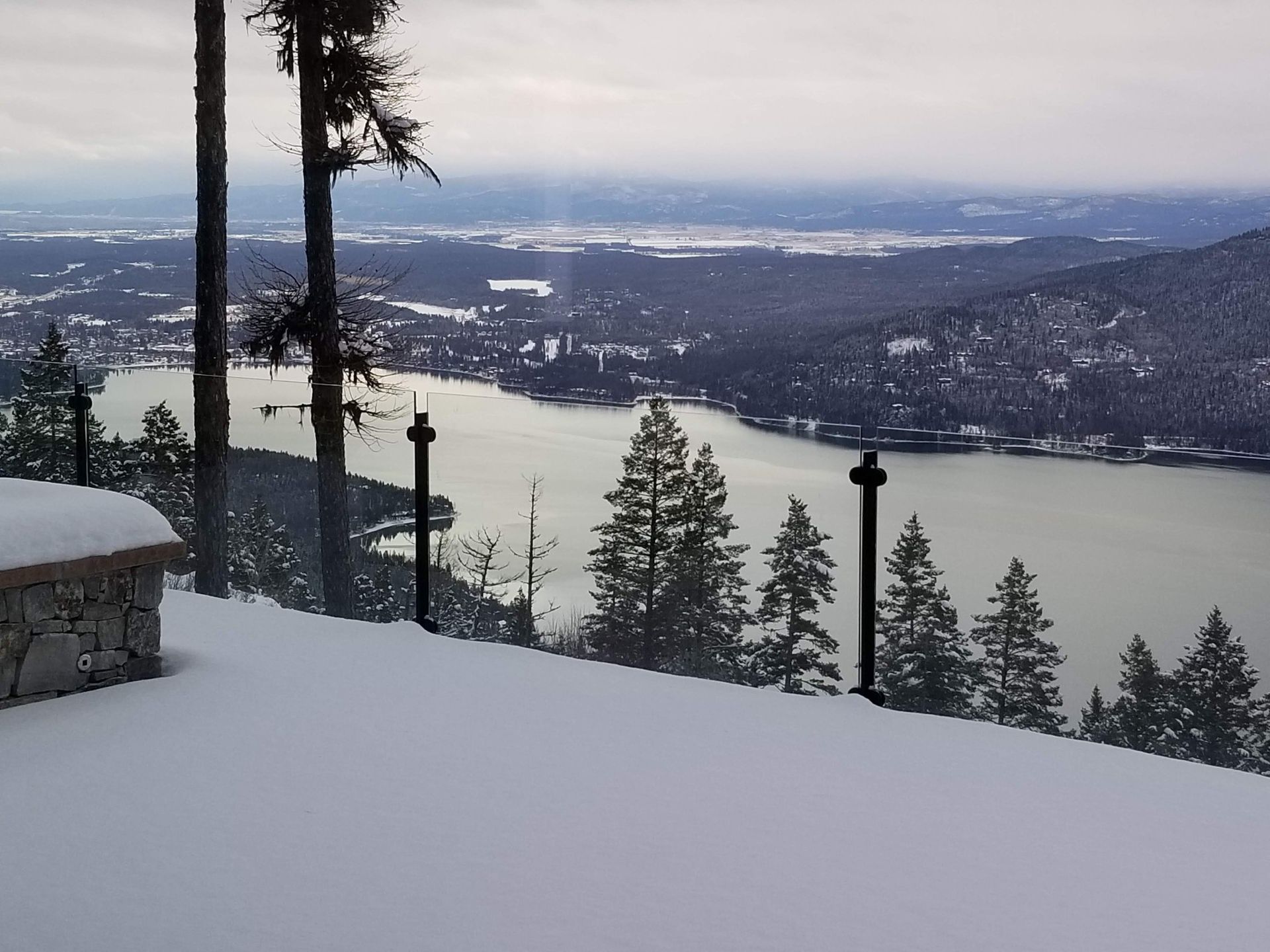 Snowy landscape overlooking a lake and town, with trees and a fence in the foreground.