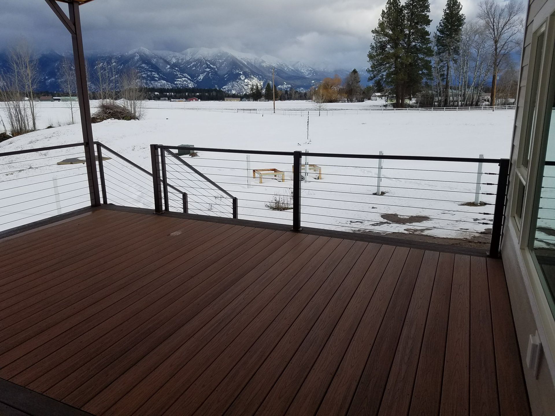 Brown deck with black railing overlooking a snowy field and mountains on a cloudy day.