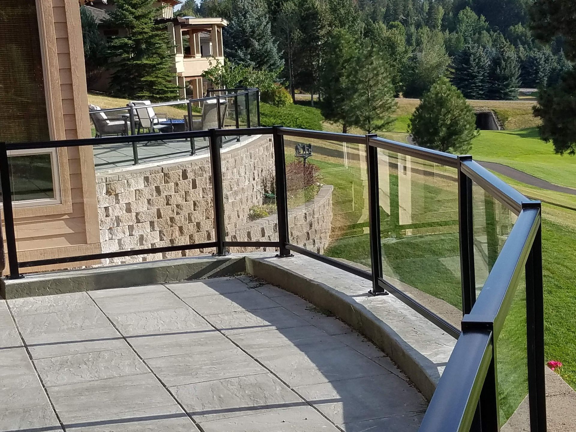 Glass railing surrounds a curved patio with a stone wall and a view of a grassy hill.