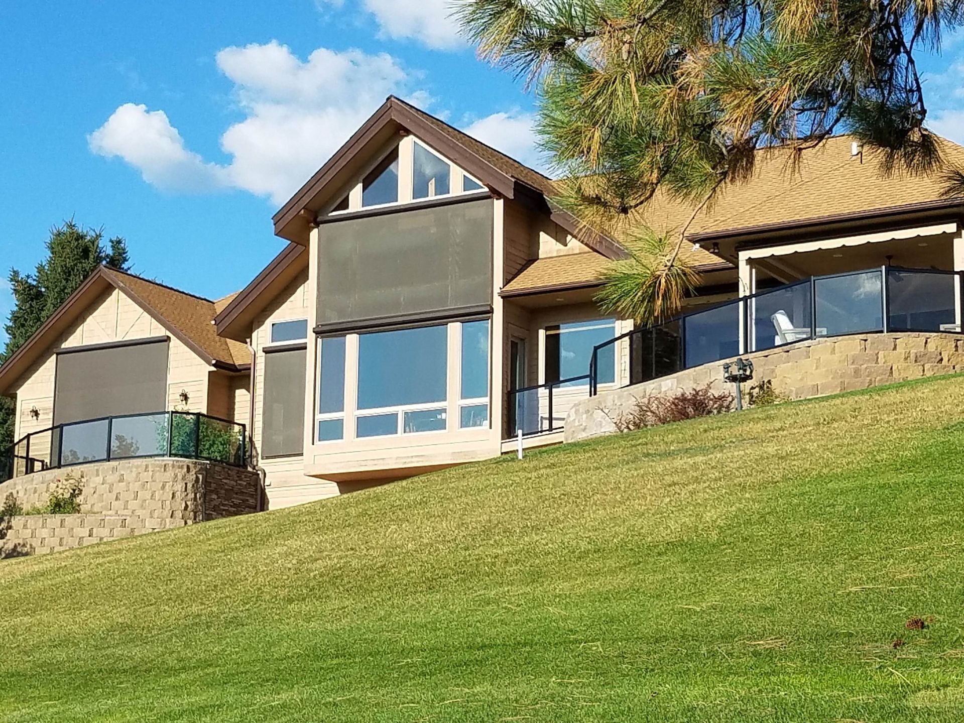House with multiple levels, brick and brown roof, glass railings, on a grassy hill under a blue sky.