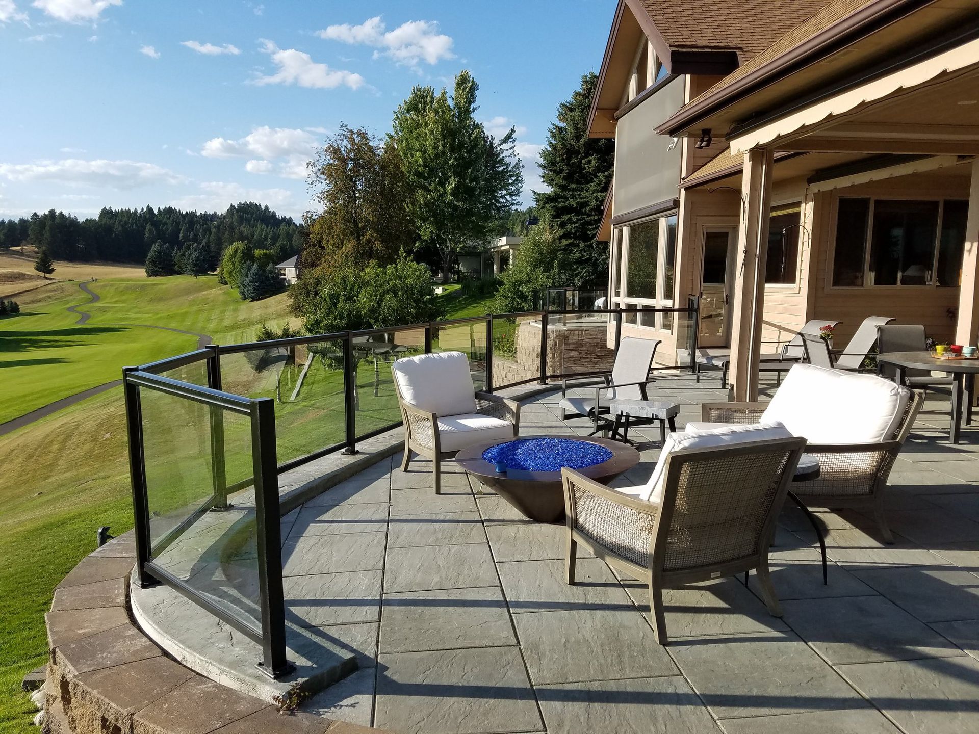 Patio with glass railing overlooking a green field. Furnishings include chairs and a fire pit.