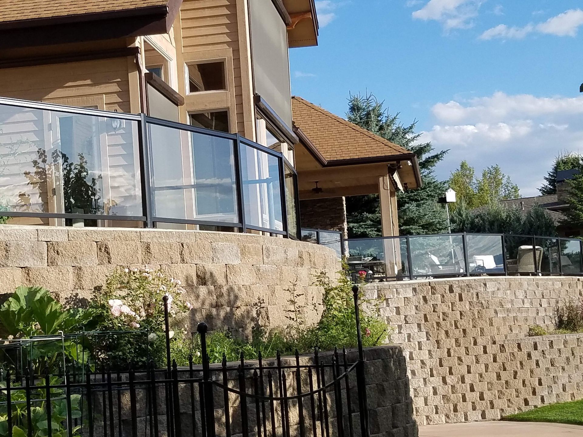 House with glass balcony railing atop a stone retaining wall. Black fence in foreground, blue sky.