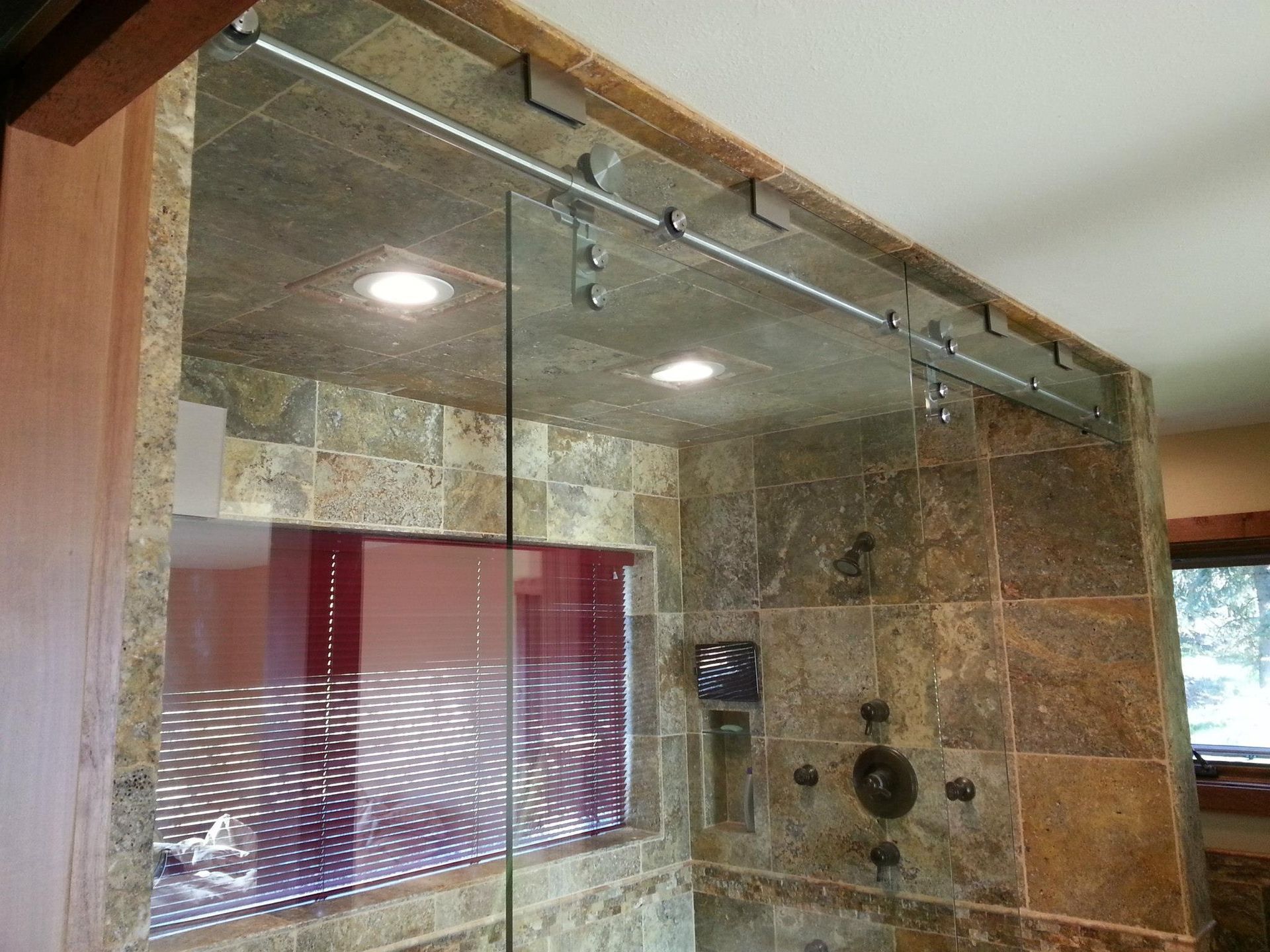 Shower with sliding glass door on a metal track, brown tiled walls, and ceiling lights.