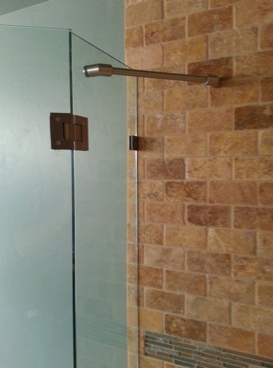 Glass shower door with bronze hinges and handle, mounted on a brick-patterned tile wall.