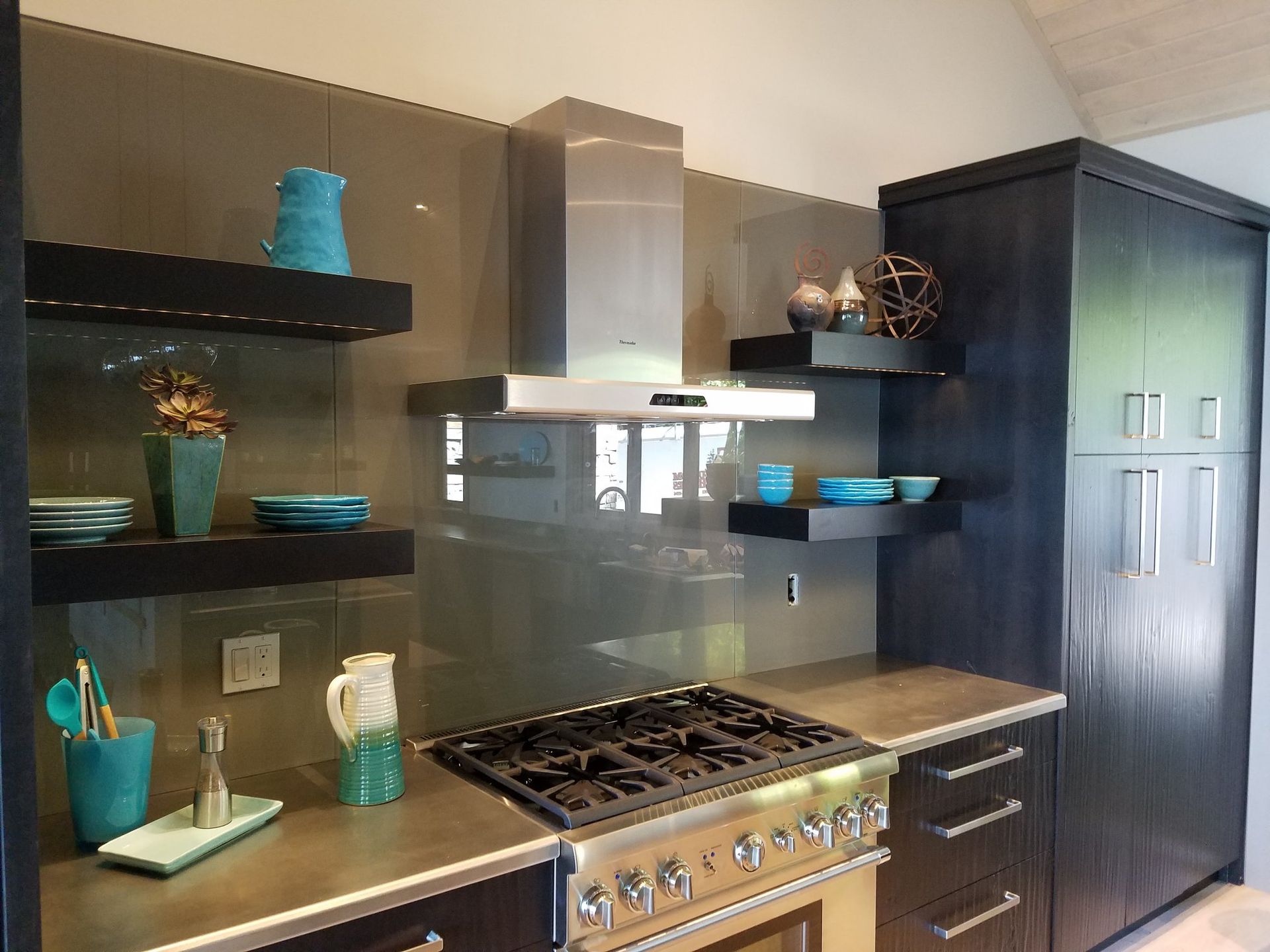 Modern kitchen with stainless steel appliances, black cabinets, teal decor, and floating shelves.