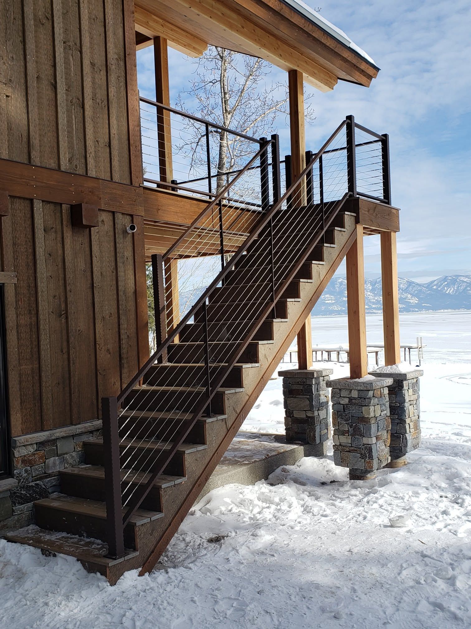 Wooden stairs with metal railing lead up to a cabin with snow and mountains in the background.