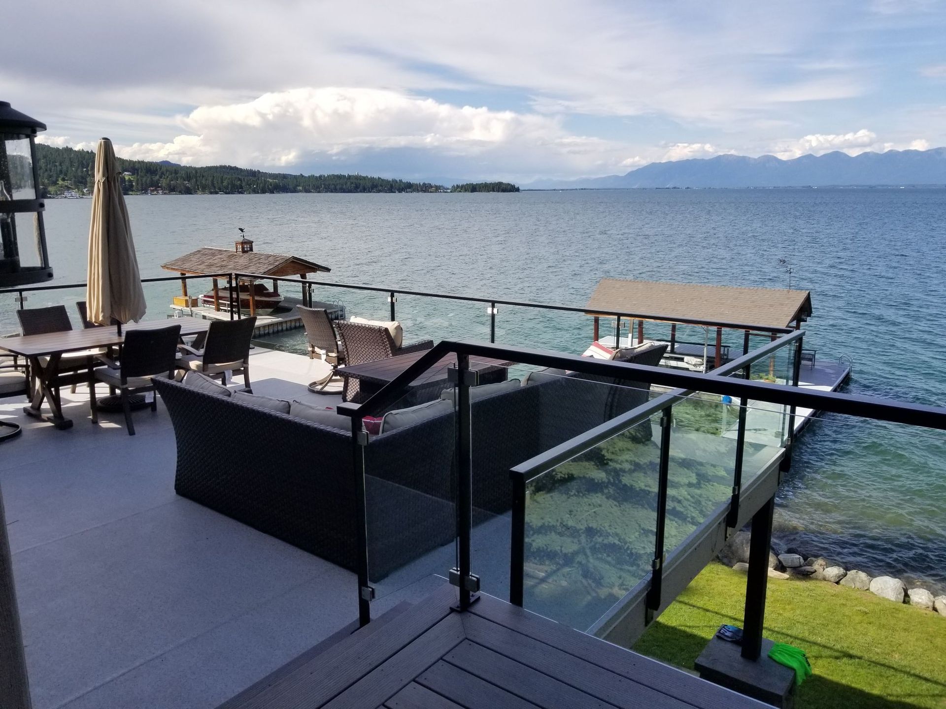 Lakefront deck with furniture, glass railings, and dock on a sunny day. Mountains in the distance.