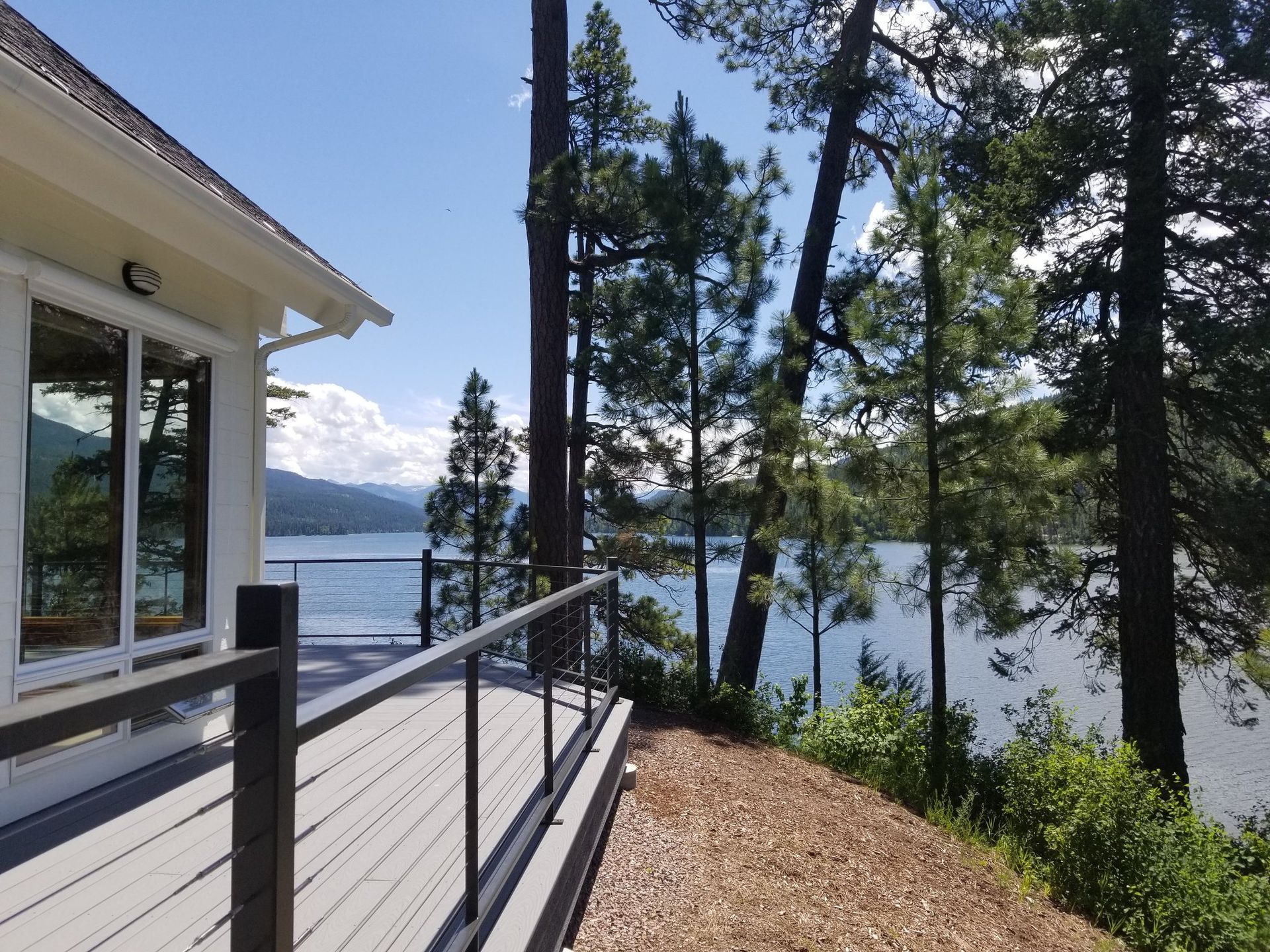 Deck overlooking a lake, with trees to the right and a house to the left, on a sunny day.