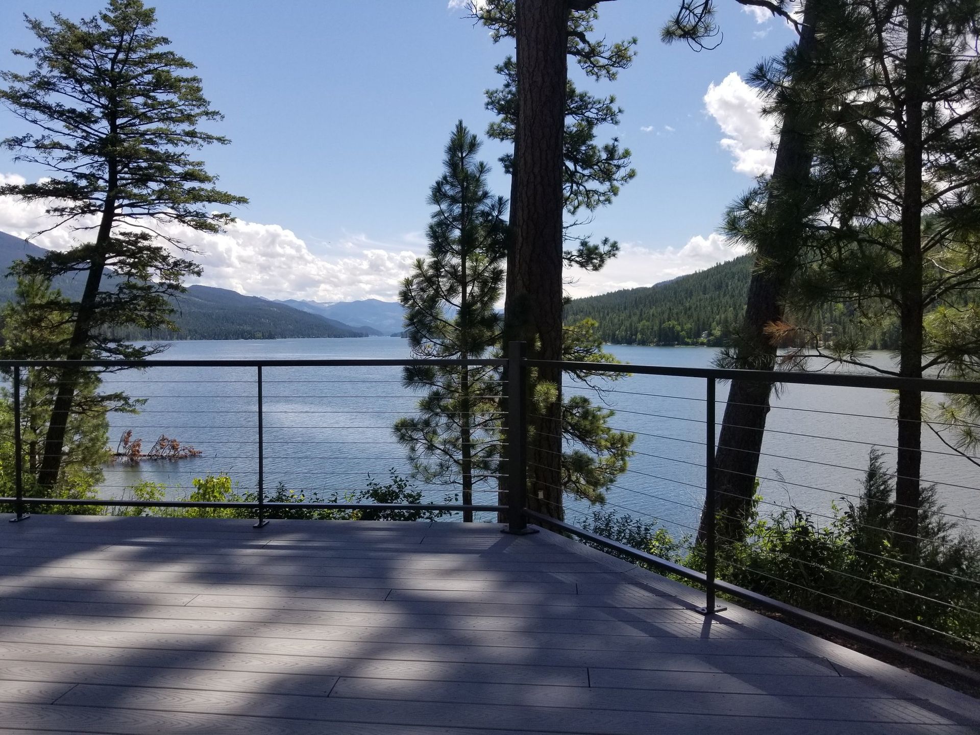 Deck overlooking lake and mountains framed by trees on a sunny day.