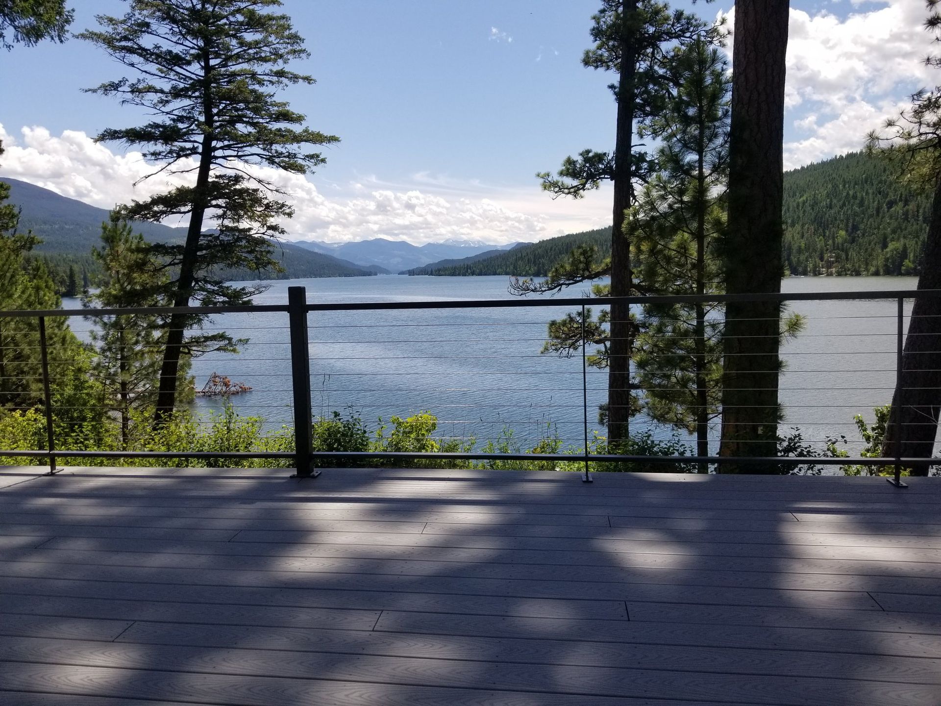 Lake view from a deck with trees and mountains in the distance, under a blue sky.