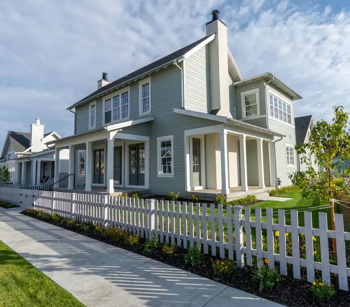 White two-story house with porch, gray siding, and a white picket fence along a sunny sidewalk.