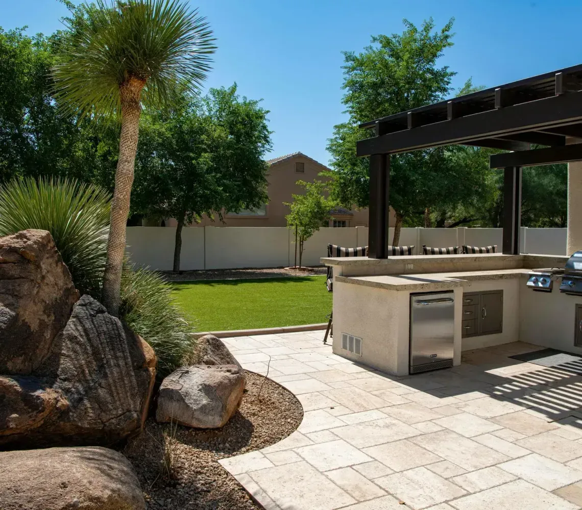 Backyard patio with stone pavers, built-in grill, pergola, and desert landscaping under a clear blue sky