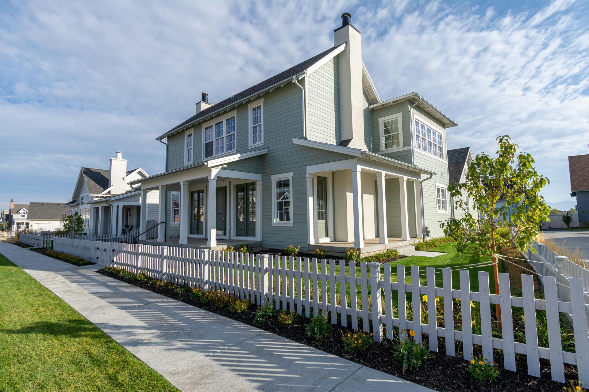 White two-story house with porch, gray siding, and a white picket fence along a sunny sidewalk.