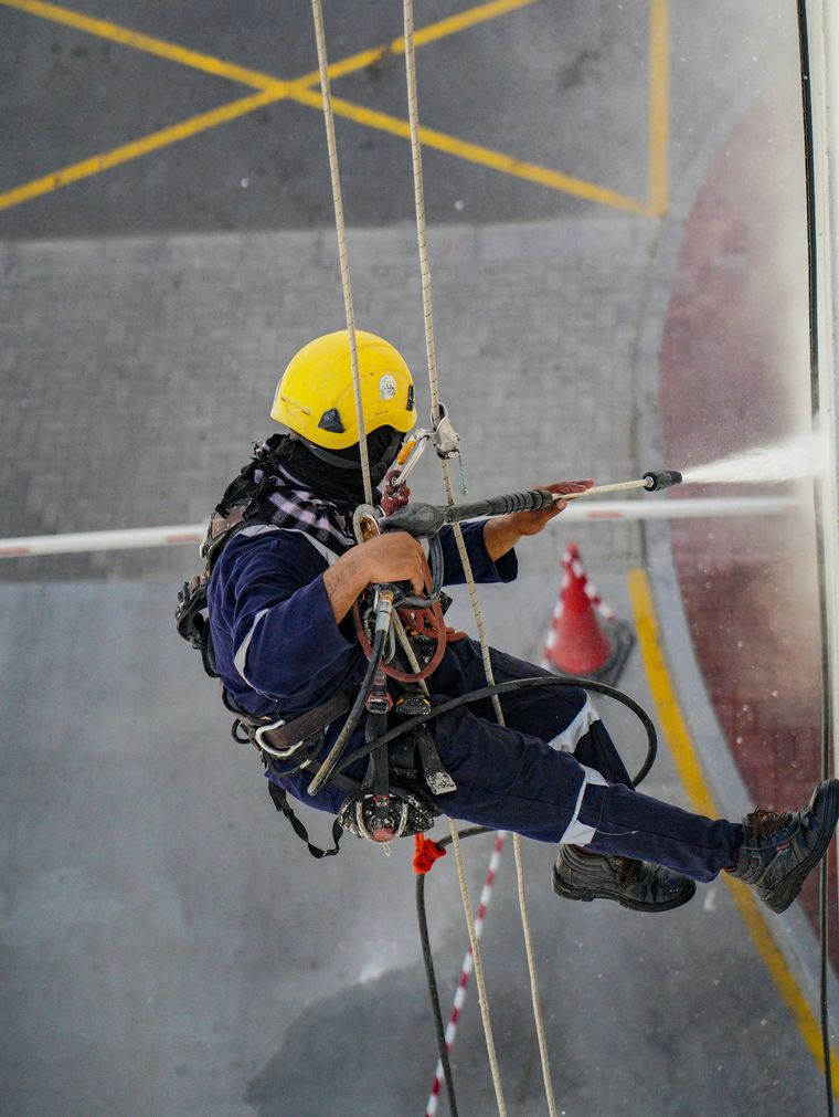 Rope-rescue worker in yellow helmet rappels down a building, spraying water at a window.