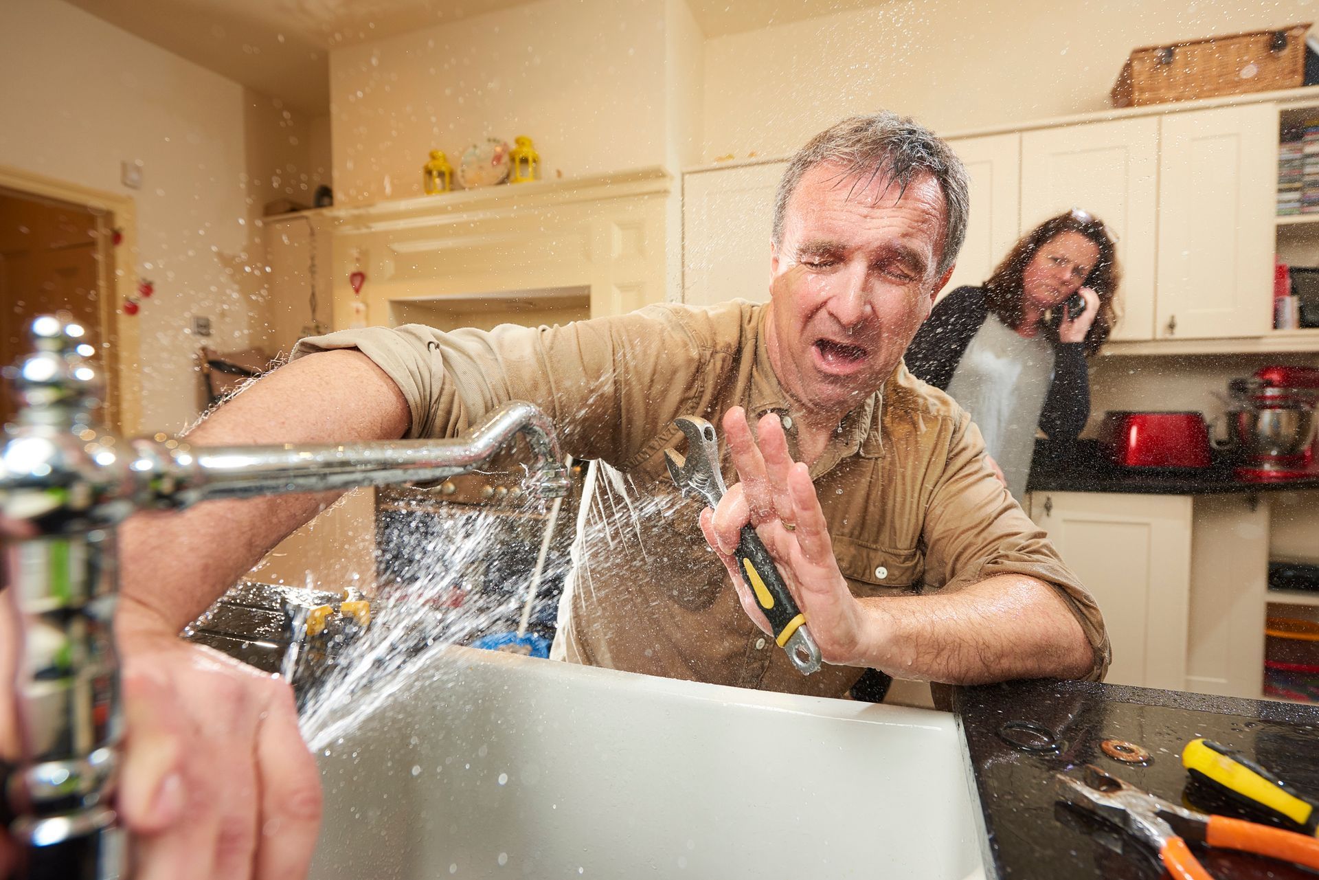 A man tries his hand at fixing the tap. A woman is already on the phone to an emergency plumber as w