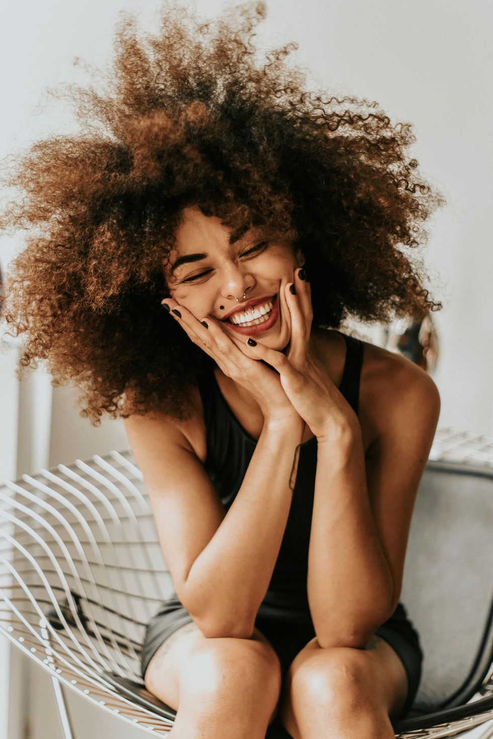 Woman with large afro smiles brightly, hands on her cheeks, sitting in a wire chair.