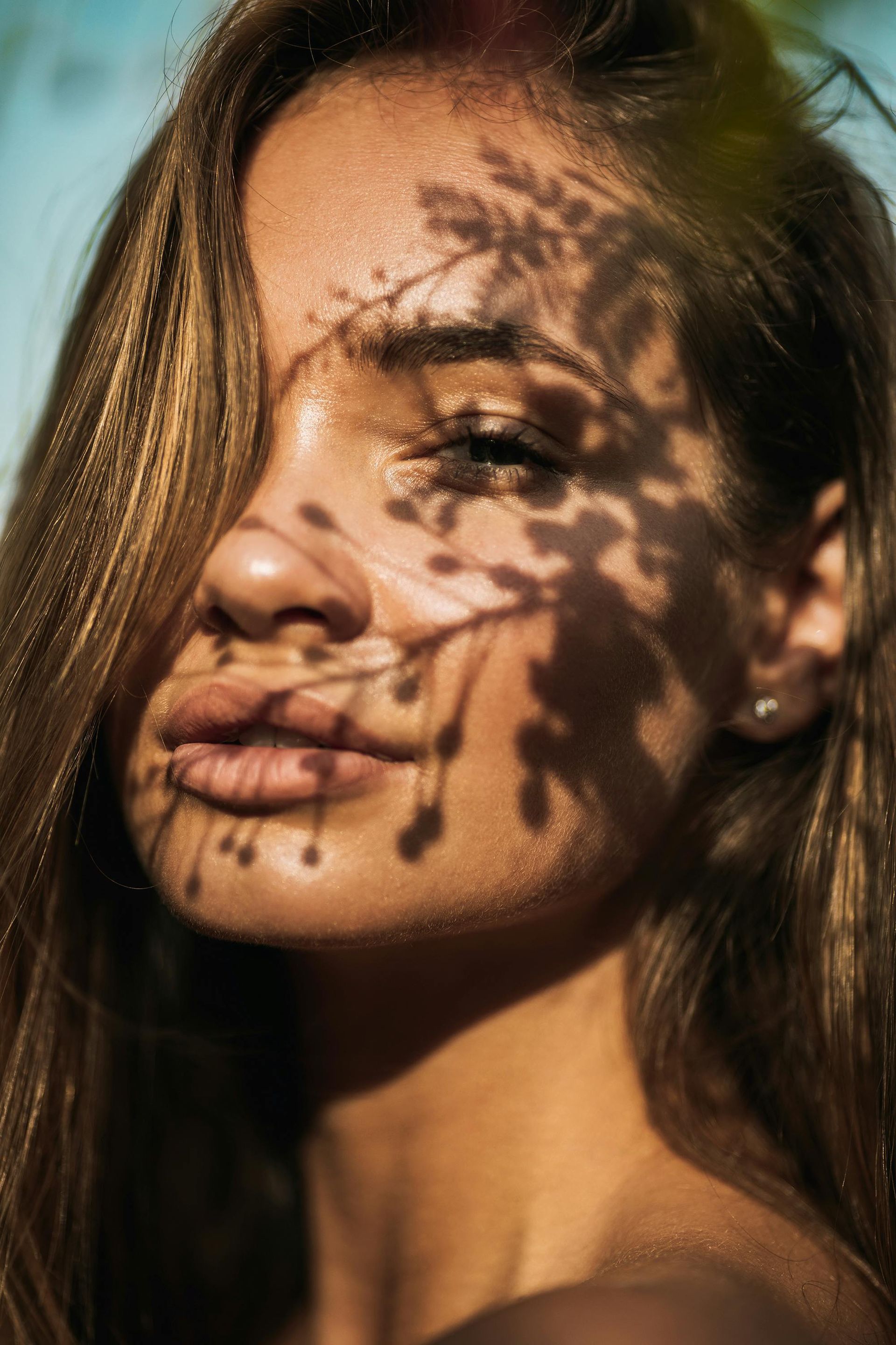 Woman with long brown hair, face in sunlight with shadow of leaves, looking towards camera.