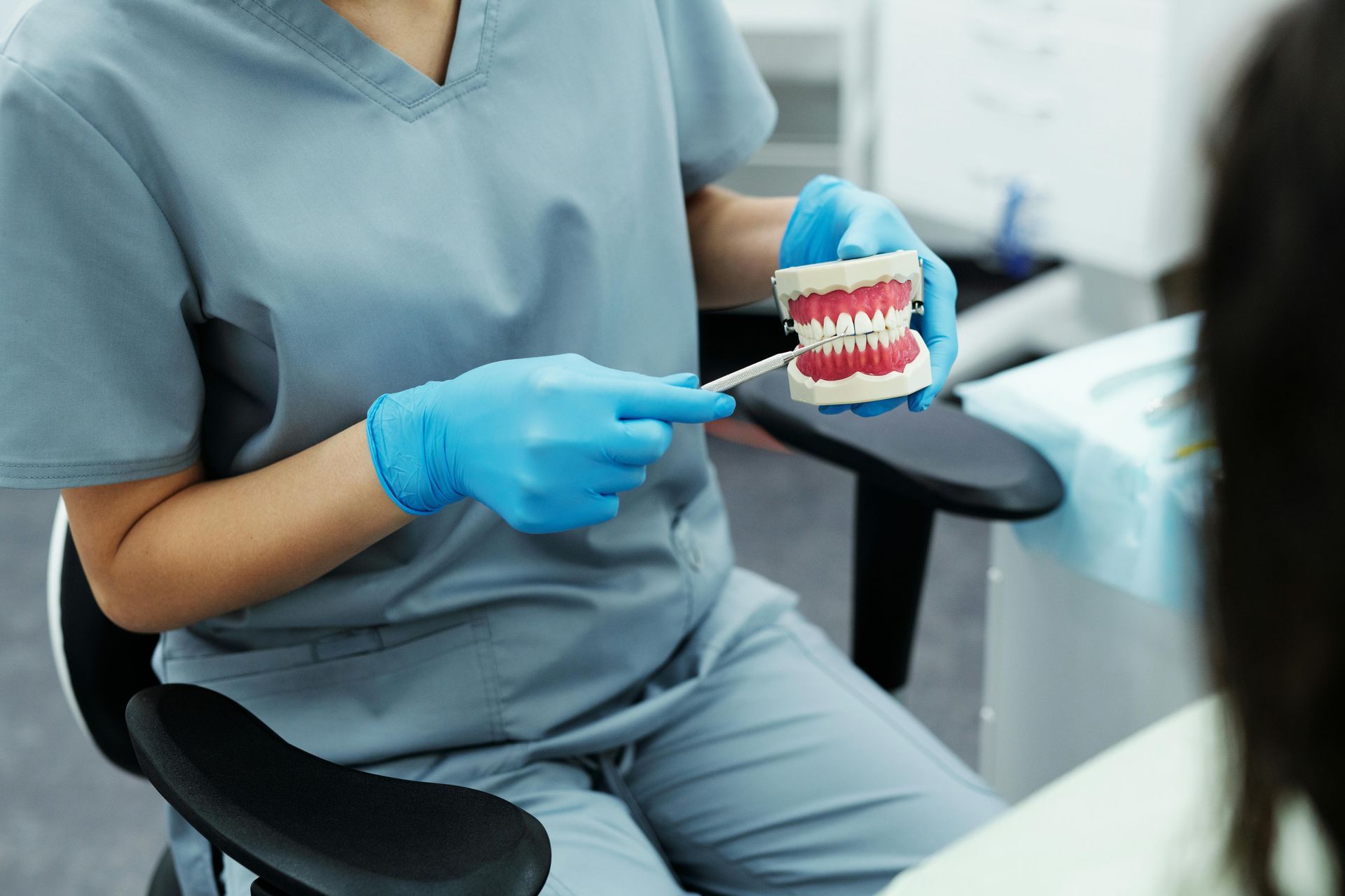 Dentist in blue scrubs and gloves pointing at a teeth model while consulting a patient.