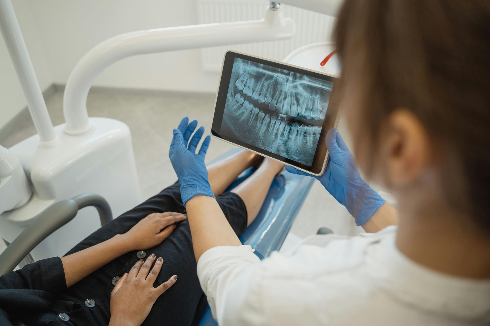 Dentist in blue gloves showing patient dental X-ray on tablet in clinic.
