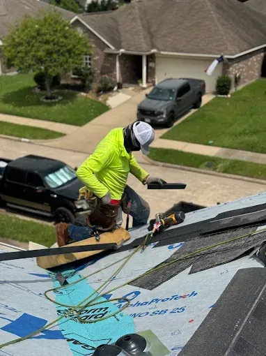 Roofer in safety gear works on a residential roof, using a power tool; a truck is parked nearby.