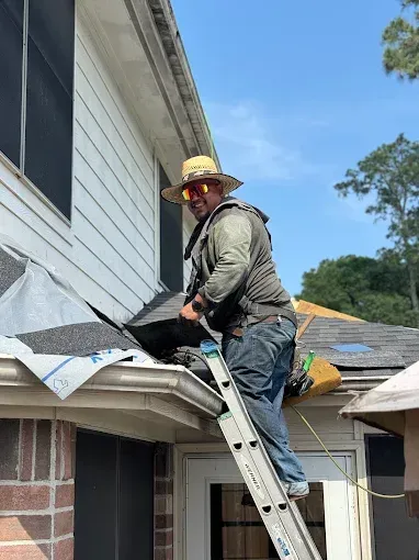 Man on ladder repairing roof, wearing straw hat and sunglasses. Outdoors, sunny day.