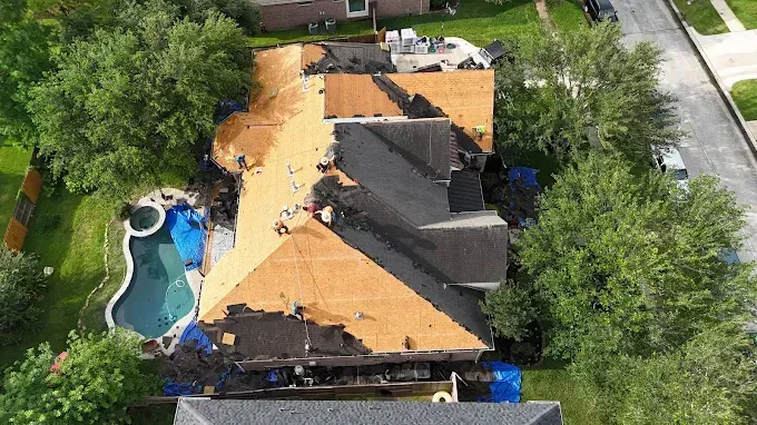 Aerial view of a house with significant roof damage and blue tarps, near a pool and street.