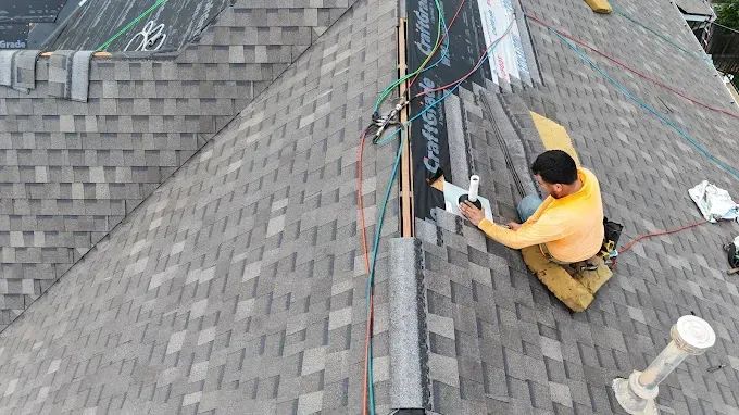 Roofer installing shingles on a roof. The roofer is using a nail gun and is wearing a yellow shirt and khaki pants.