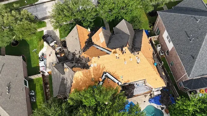 Aerial view of a house with significant roof damage and workers present. Construction underway, near a pool.