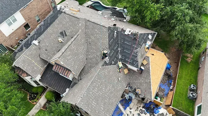 House with roof damage, under repair, asphalt shingles, workmen on the roof, debris below, green foliage and pool nearby.