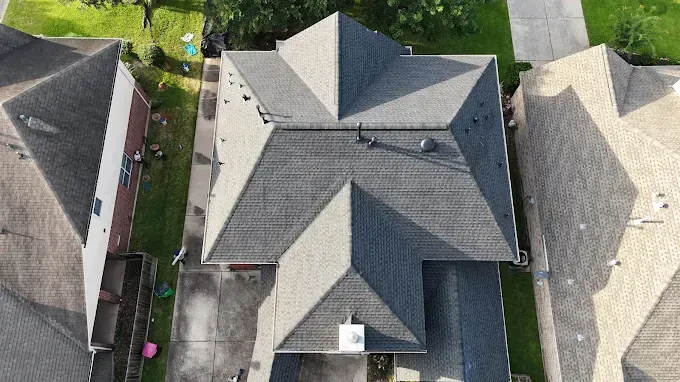 Overhead view of a house with a complex, dark gray roof, surrounded by other houses and pavement.