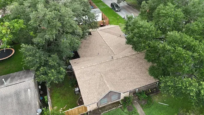 Aerial view of a house with a brown roof surrounded by green trees and lawn; a street and other houses are visible.