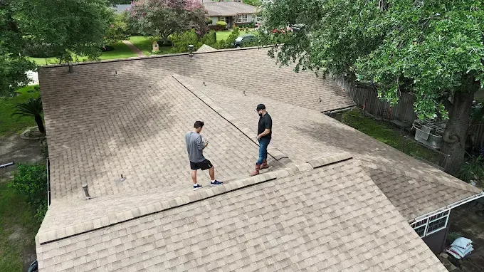 Two people on a brown shingle roof inspecting it near trees and houses.