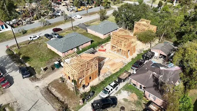 Aerial view of houses under construction with exposed wooden frames, surrounded by other homes and cars.