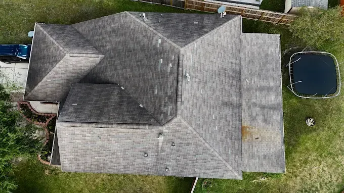 Overhead view of a house with a gray roof, surrounded by green grass. A small black car sits to the left.
