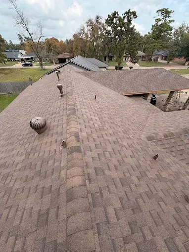 Brown asphalt shingle roof on a house, with vents and a background of trees and houses.