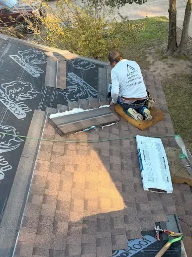 Roofer on a roof installing shingles. Beige shingles, black underlayment, and a white container are visible.