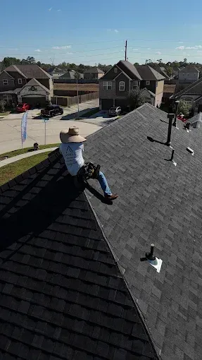 Roofer in straw hat sits on a dark shingled roof, working. Houses and blue sky in background.