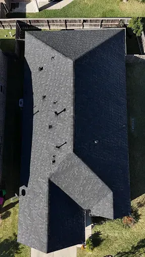 Overhead view of a dark gray asphalt shingle roof on a house with vents, surrounded by grass and other buildings.