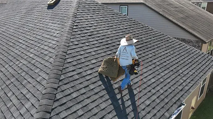 Person on a dark shingle roof, holding a large bundle. Blue jeans, white hat, sunny.