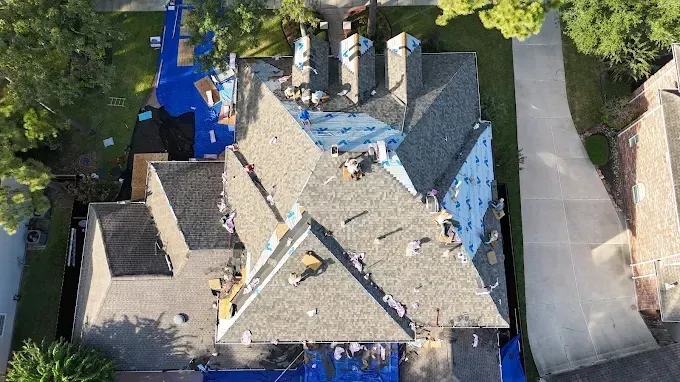 Overhead view of a house roof undergoing repair. Workers, tarps, and materials visible on the gray shingled roof.