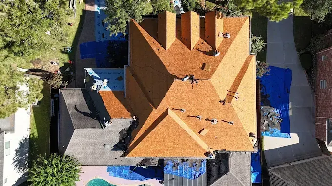 Aerial view of a house with orange shingles partially replaced and covered with blue tarps.