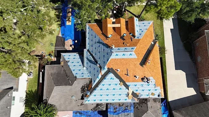 Aerial view of a house roof partially covered with brown shingles and blue underlayment, under construction.