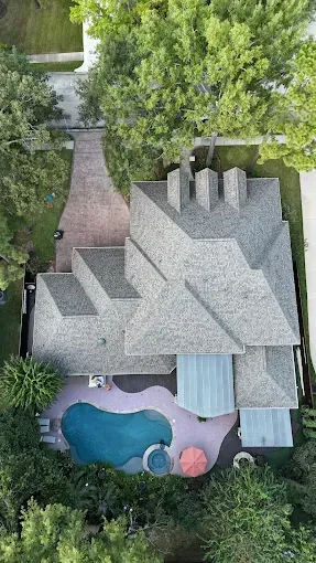Aerial view of a house with a pool and brick driveway, surrounded by trees.